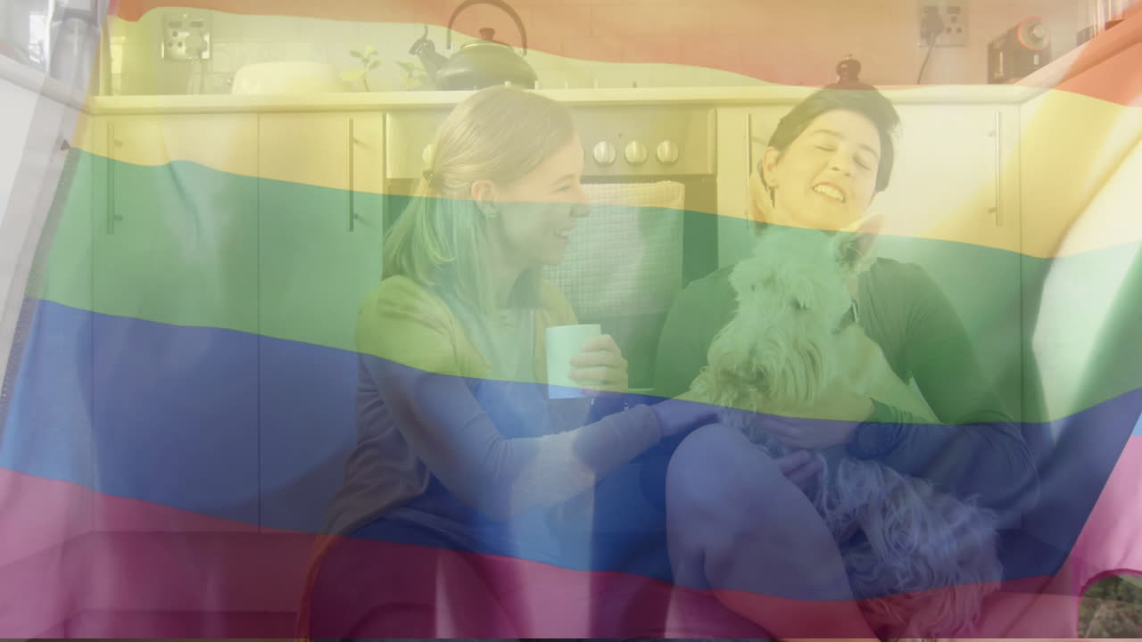 animación de la bandera del arco iris del orgullo sobre una feliz pareja de lesbianas caucásicas sentadas con un perro mascota en la cocina