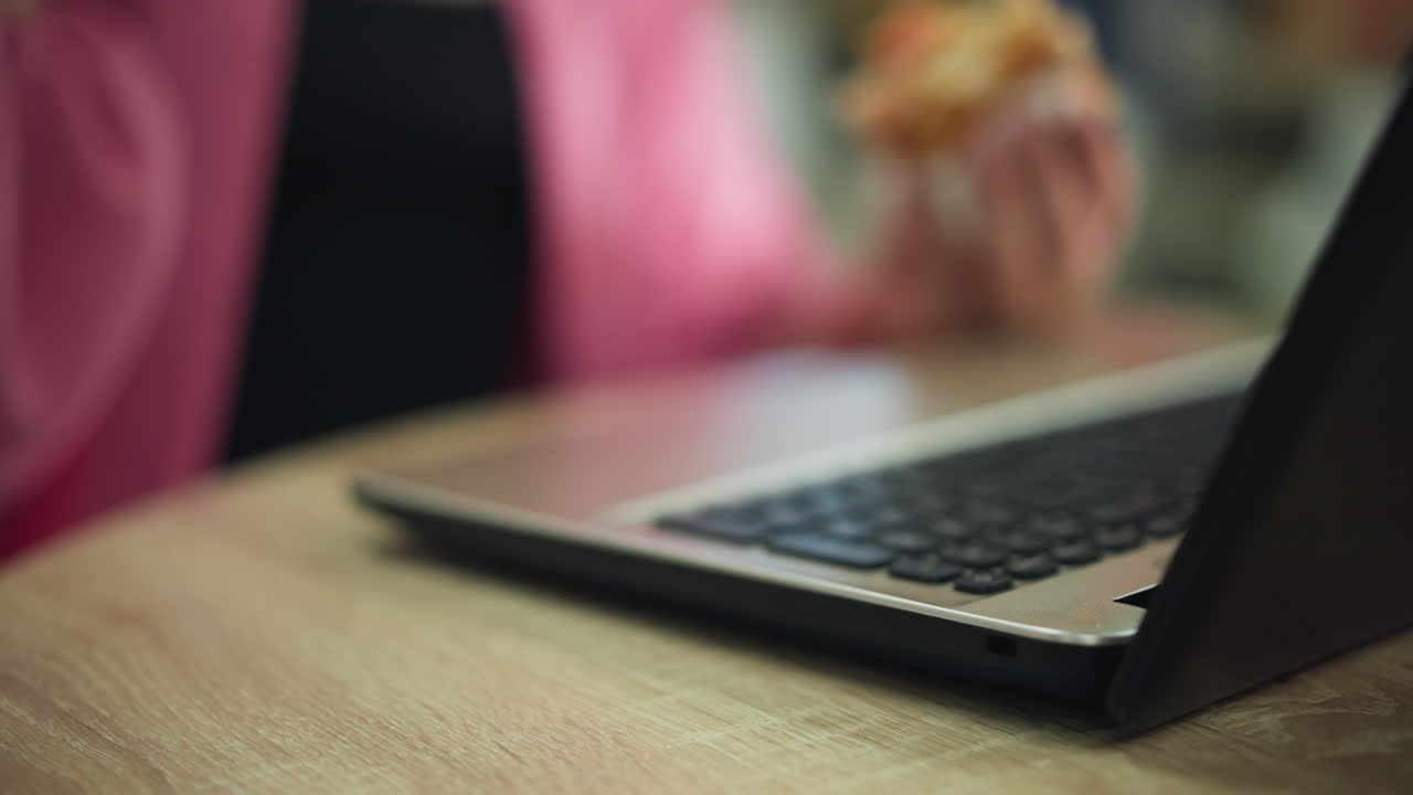 나무 테이블 위에 있는 노트북의 클로즈업, 한 손에 햄버거를 들고 다른 손에 커피 컵을 들고 커피를 테이블에 다시 부드럽게 놓고, 배경이 흐릿합니다.