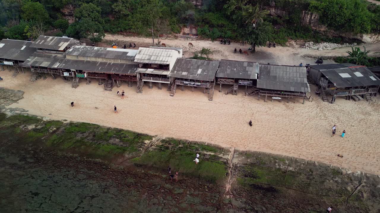 Cinematic drone footage shows an Indonesian shoreline community with timber beachfront shacks, coral reef at low water, calm ocean and white sand beach shaped by steady coastal movement