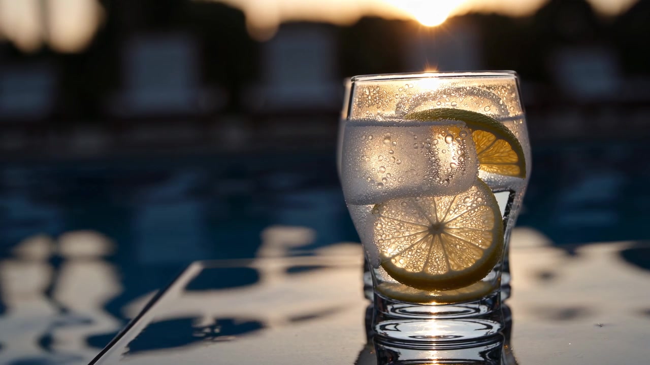 Bicchiere rinfrescante di acqua al limone con ghiaccio a bordo piscina al tramonto