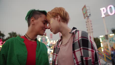 Close-up-shot-of-a-girl-in-a-checkered-pink-shirt-and-a-girl-in-a-green-shirt-holding-hands-and-bringing-their-heads-together-and-rubbing-their-foreheads-together-during-their-warm-date-in-the-amusement-park