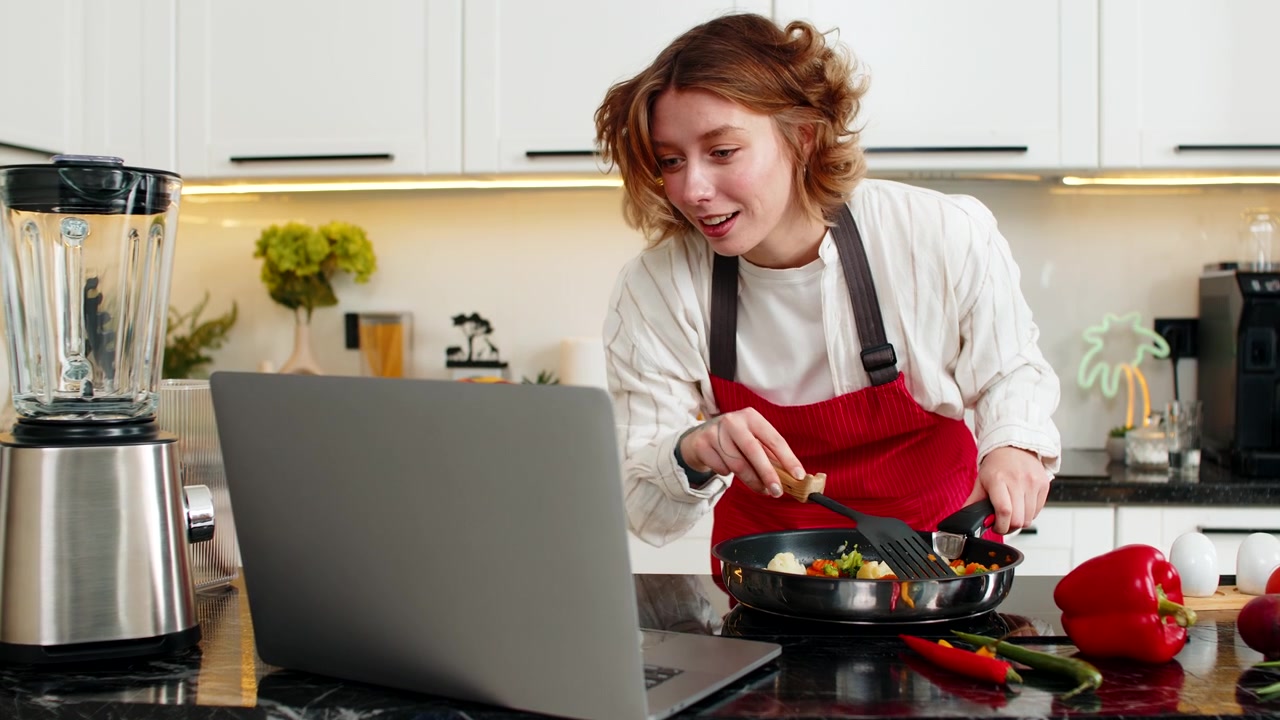 Ung kvinde laver grøntsags-wok, mens hun ser en opskriftsvideo på laptop til et diætmåltid i hjemmekøkkenet.