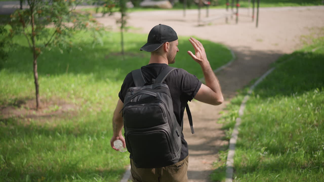 Man Waves Goodbye, Person Departs With Friendly Gesture, Man Making Casual Gesture As He Walks Through Park, Male Figure Waving Farewell While Strolling Along Sunny Park Path With Backpack