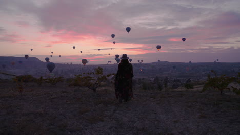 lady in dress walking outdoor to view stunning hot air balloons scenery in the sky at dawn in cappadocia, turkey, wide, following shot