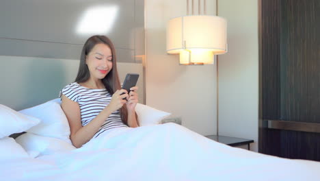 a cute young woman, sitting up in a big comfy bed relaxing as she texts on her smartphone