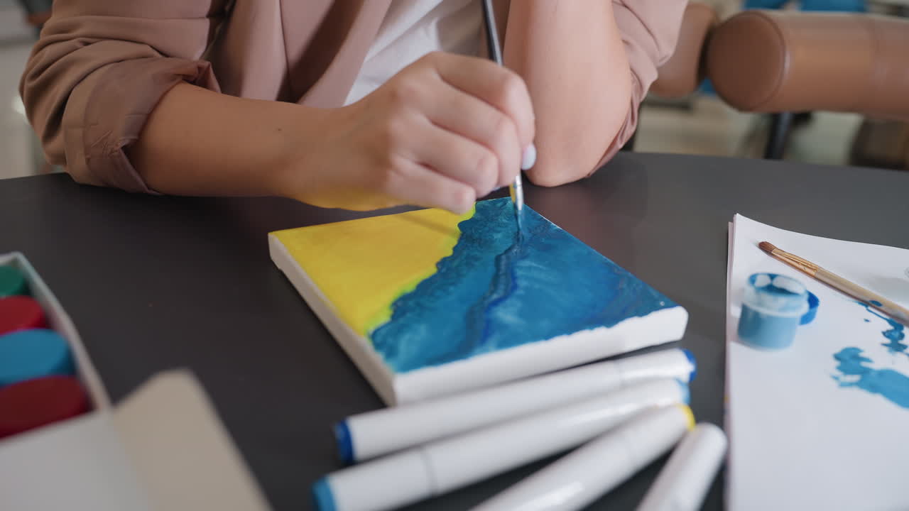 Primer plano de un pincel dando los últimos toques a los bordes azules de un paisaje marino en un pequeño lienzo, con rotuladores y un bote de pintura cerca, suavizando y acentuando con cuidado, momento íntimo de acabado en el estudio.