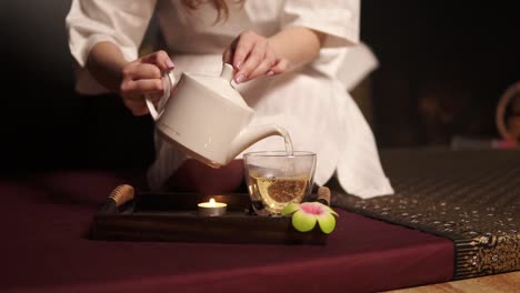 beautiful brunette in bathrobe taking teapot and pouring tea in glass cup drinking herbal tea at the spa in slow motion. tea