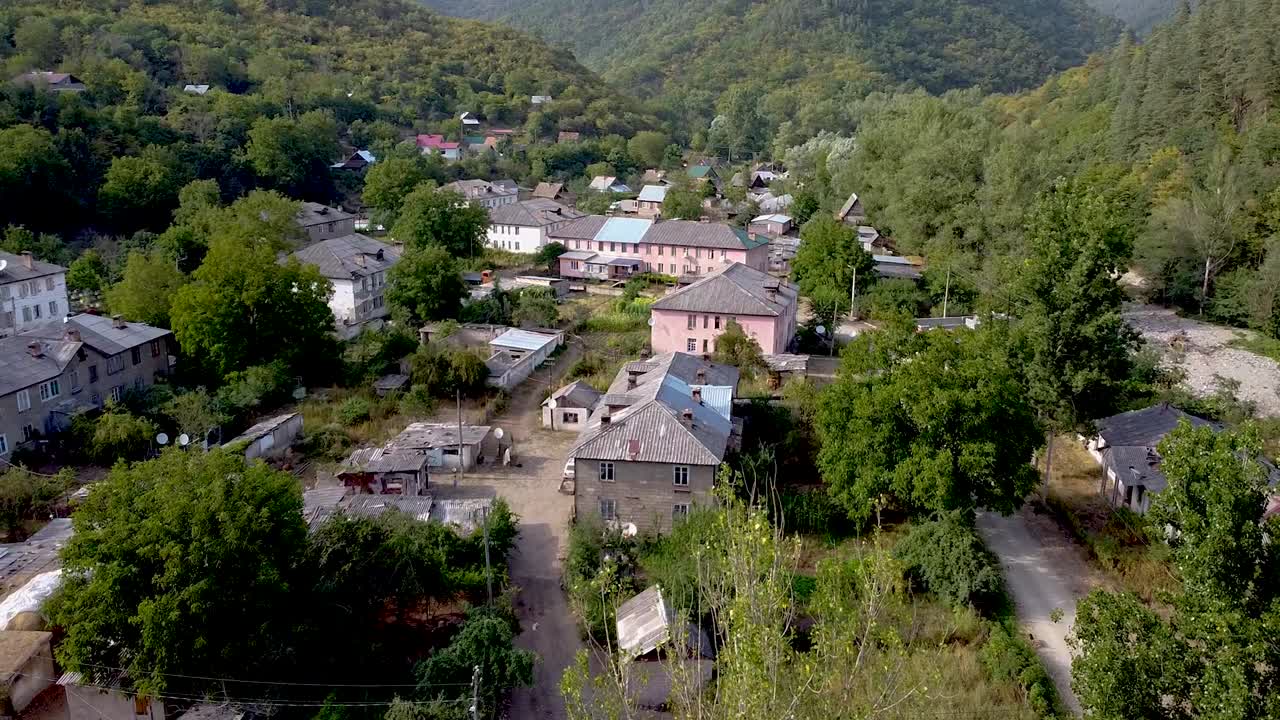 A quiet village sits nestled in the mountains, bathed in soft morning light. Houses with varied rooftops contrast against the lush trees and open fields nearby, creating a serene landscape