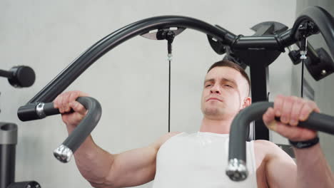 young man working out on chest press machine hands gripping curved handles eyes closed face tightened muscles straining under gym lighting on sleek black steel equipment in modern gym facility