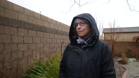 eine schöne alte frau mit brille, die in einem regensturm läuft und einen regenmantel und eine kapuze trägt, während regentropfen in zeitlupe fallen