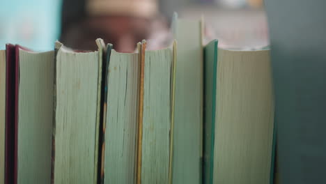 tight macro view of books on shelf as person reaches to take one, edges and spines filling frame, soft library bokeh behind, moment of choice during quiet study search