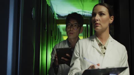 Diverse-female-and-male-it-technicians-in-lab-coats-using-tablet-and-laptop-checking-computer-server