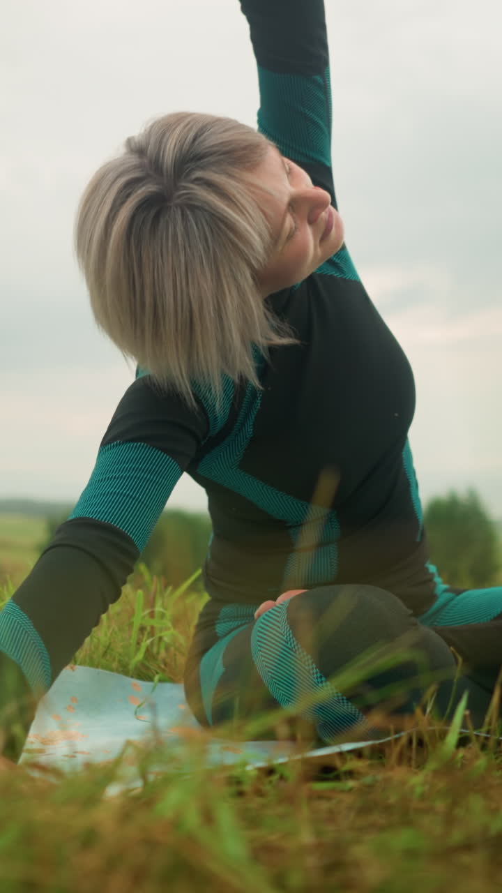 mujer de cabello gris acostada de un lado en la alfombra de yoga practicando la postura de inclinación lateral, brazos extendidos, ojos cerrados, se levanta lentamente, bajo un cielo nublado en un campo de hierba sereno con árboles en la distancia