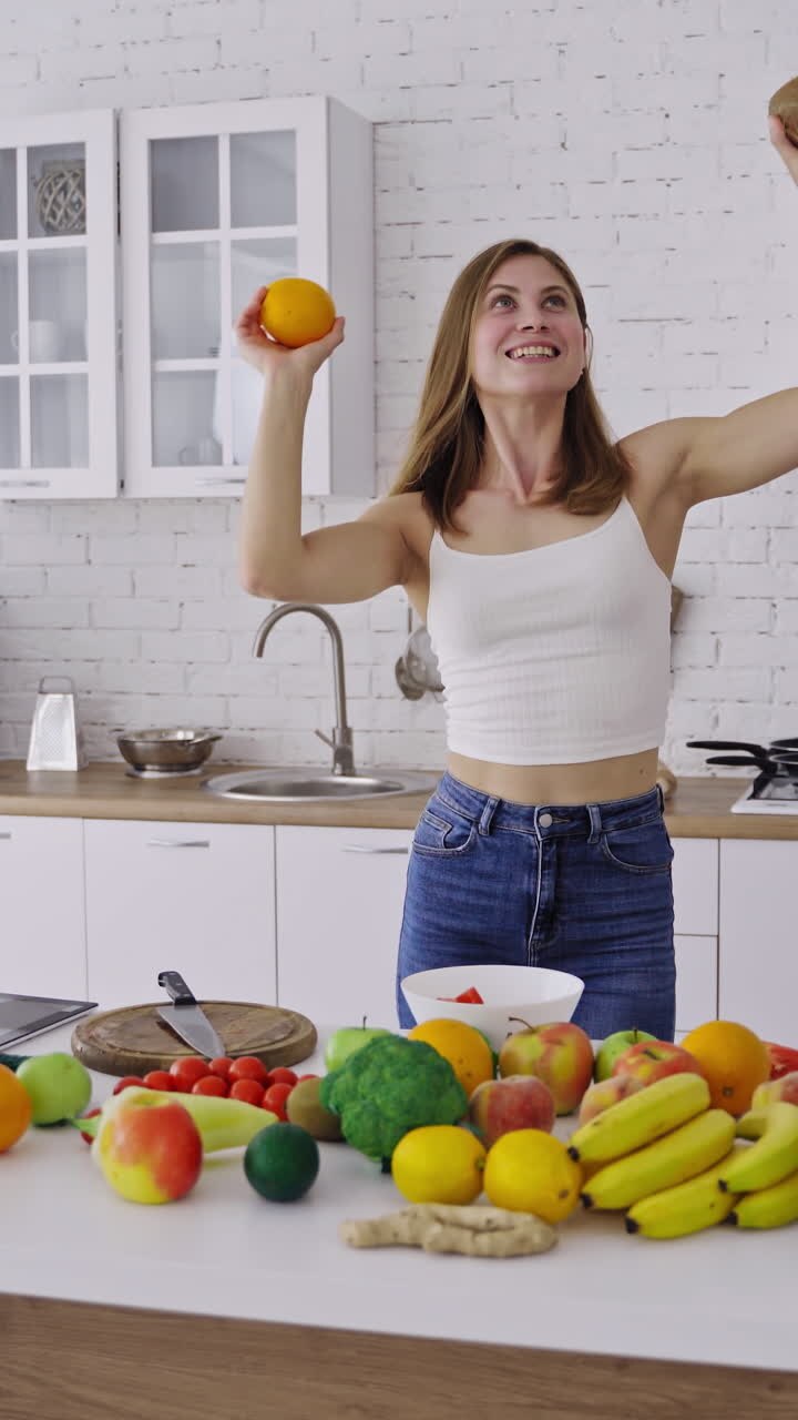 Beautiful slim woman is dancing with fresh fruits in the modern kitchen. Happy woman holding coconut and orange having fun at home. Organic fruit and vegetables. Dieting concept. Vertical video