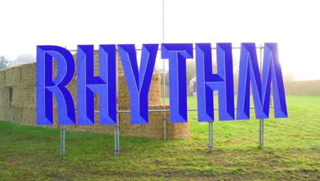 3d sign rhythm outdoor on festival designating a certain music area camera movement pull back revealing the full text cinematic atmosphere grassy field and plenty bales in the background stacked