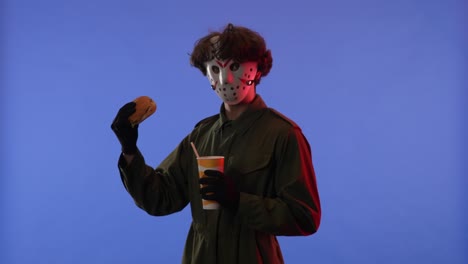 man in white mask with coke and burger in hands on blue background with red neon light image of
