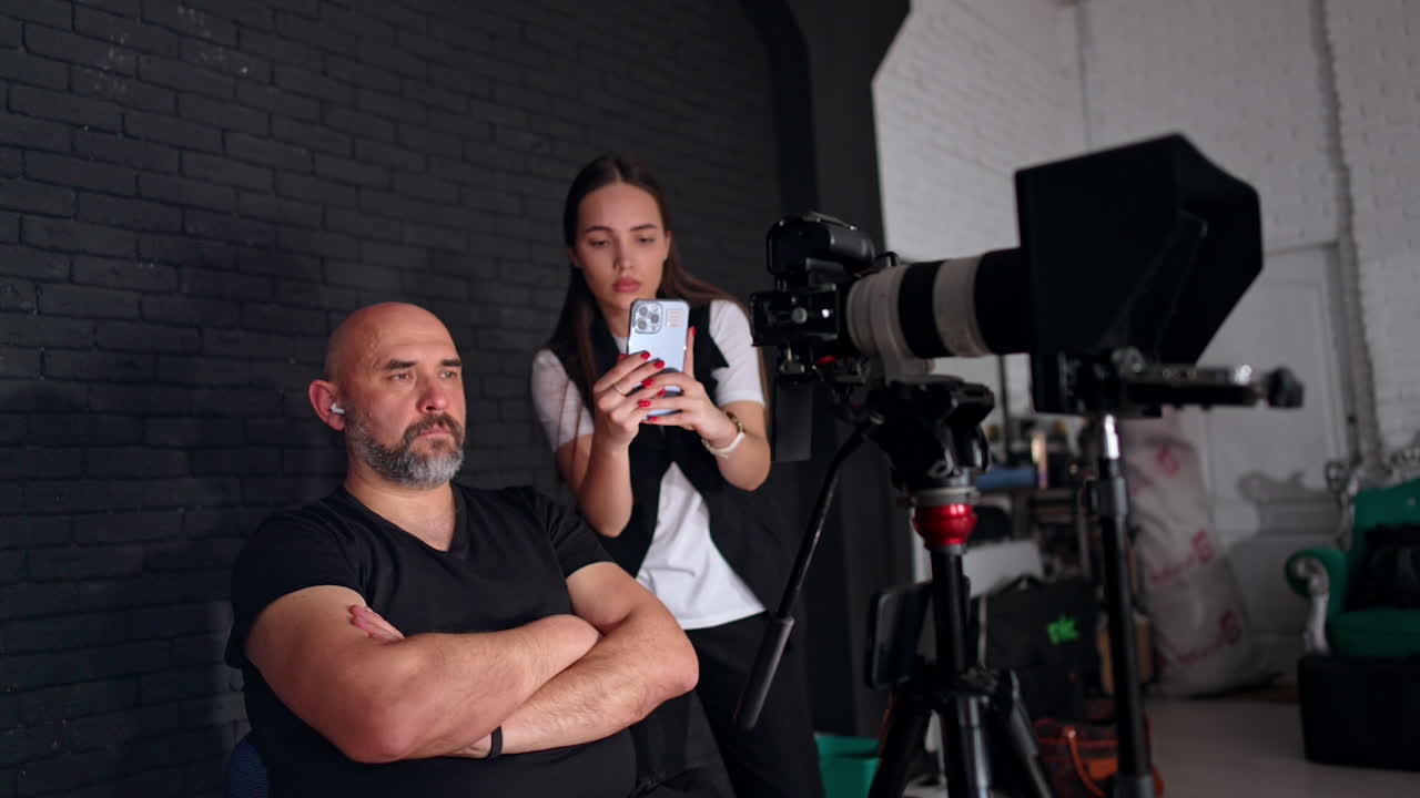 Bearded man sits with hands crossed on his chest behind the camera. Lady standing beside taking video on smartphone.