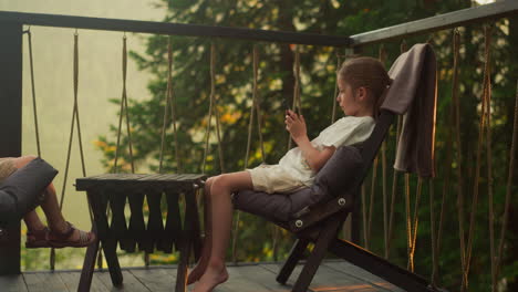 cute girl attentively watches videos alone sitting on balcony among dense forest. child concentrates on contemporary device in calm atmosphere at sunset