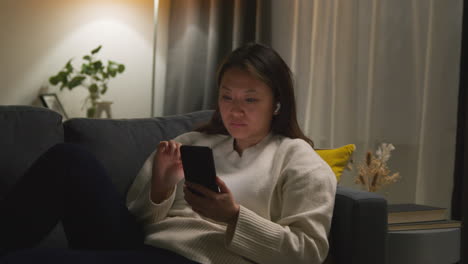 woman spending evening at home on sofa wearing wireless earbuds and streaming music or podcast from mobile phone 5