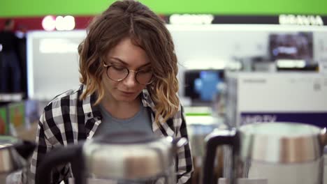 joven alegre mujer positiva en camisa a cuadros y gafas eligiendo hervidor eléctrico en la tienda de electrodomésticos, caminando en un