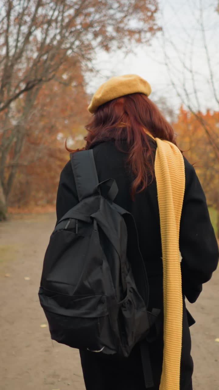 vista trasera de la dama con el cabello rizado rojo caminando por el camino del bosque rodeado de follaje de otoño, llevando una bolsa negra y usando una boina amarilla