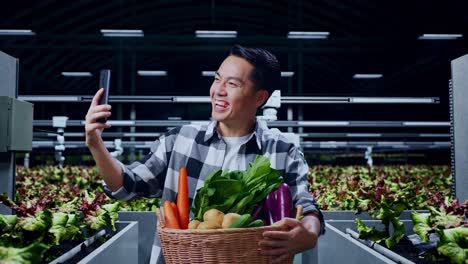 un granjero asiático con una canasta de verduras tiene una llamada de videoconferencia con alguien a través de un teléfono inteligente en una granja inteligente