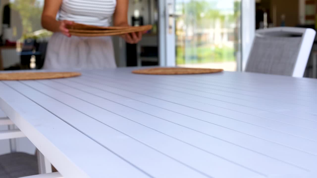 A person arranges woven placemats on a white table in a bright outdoor setting, creating a welcoming atmosphere