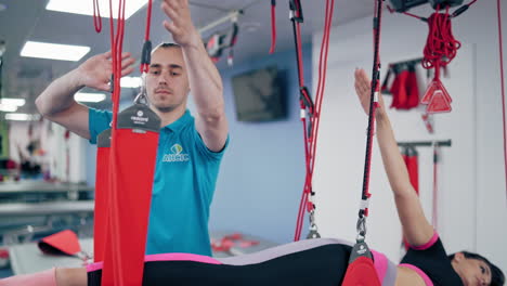 handsome male trainer helps young woman to do exercises in the rehab center. young man physiotherapist controls the working out of a woman with special rope system indoors.