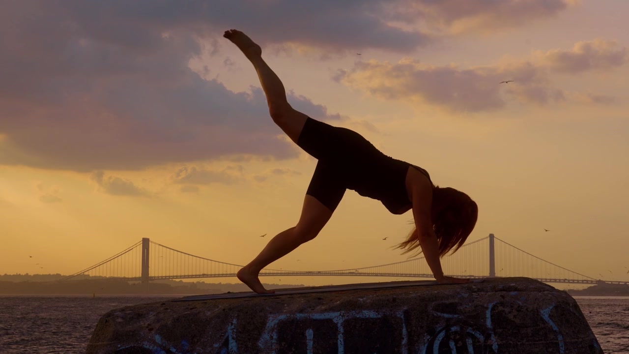 Donna in posizione plank su un molo di pietra vicino a un ponte di New York. Silhouette femminile a figura intera di profilo che pratica un esercizio yoga di flessione in riva al mare di fronte al ponte Verrazzano.