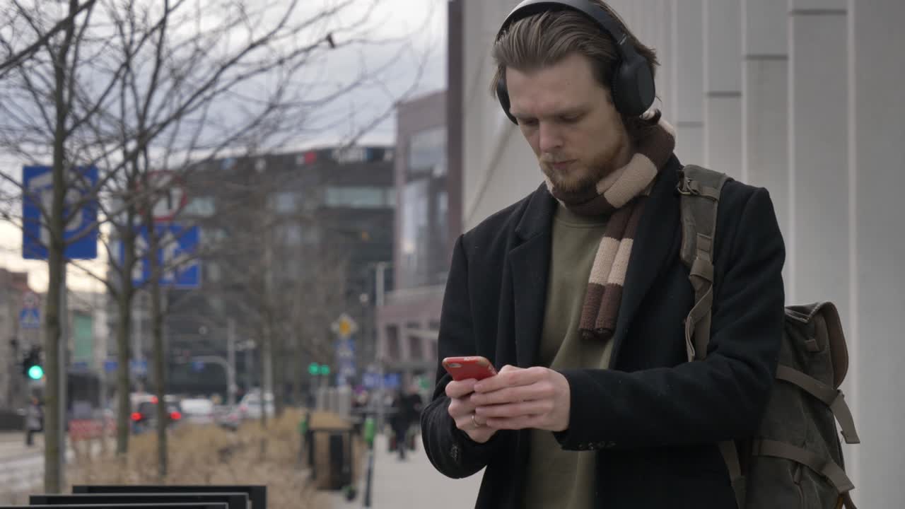 스카프와 코트를 입은 세련된 남자가 휴대전화를 사용하여 스트리밍 서비스에서 음악을 듣습니다.