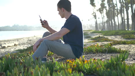 Ein-Junger-Kaukasischer-Mann-Sitzt-Am-Strand-Und-Schaut-Ruhig-Auf-Sein-Smartphone-In-Santa-Barbara,-Kalifornien