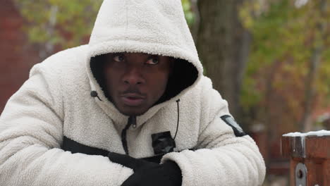 rap artist in white hoodie and gloves leans on iron bar outdoors, nodding his head in thought, background features snowy ground, trees, and distant buildings