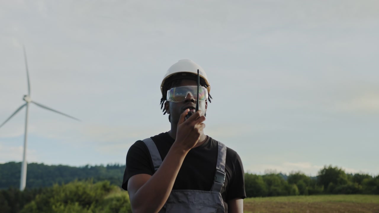 Afroamerikansk vindmølleingeniør iført hat og sikkerhedsbriller holder radio nær vindmølle