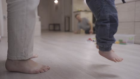 baby sitting on the floor looking at camera. his mother helps him get up and take his first steps while his father waiting for him in the kitchen playing with a toy truck