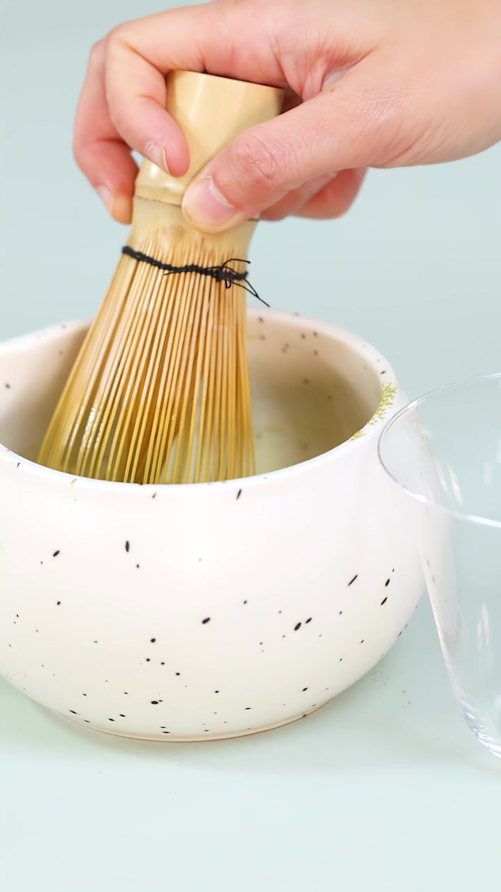 Primo piano di una mano che sbatte il tè verde matcha in una ciotola di ceramica maculata, con illuminazione morbida da studio.