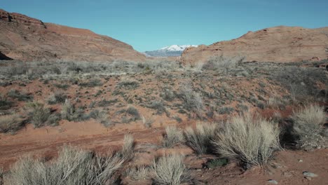 Niedriger-Drohnenflug-über-Die-Karge-Landschaft-Utahs-Mit-Blick-Auf-Die-Schneebedeckten-Berge-In-Der-Ferne