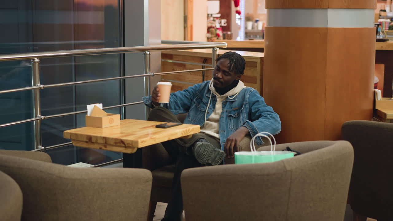 Guy in denim jacket and beige hoodie sits cross-legged in cozy caf chair, holding coffee cup in hand, smartphone on wooden table, shopping bag nearby, peaceful moment in warm modern indoor setting