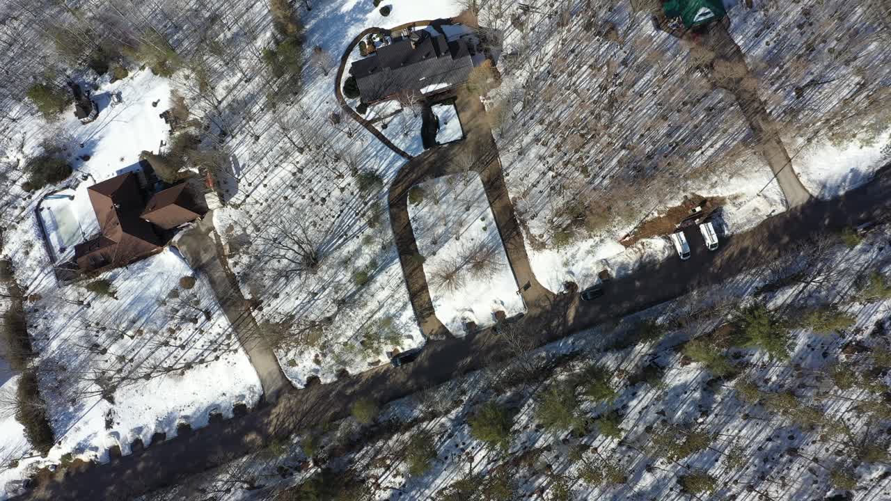 High angle aerial perspective slowly panning over suburban houses with long driveways, surrounded by a snow covered forest during a sunny winter day, showcasing the quiet rural landscape