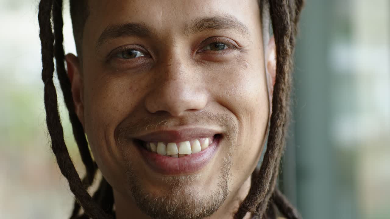 Homem de meia-idade com dreadlocks passando de uma expressão séria para um largo sorriso em frente a uma vidraça