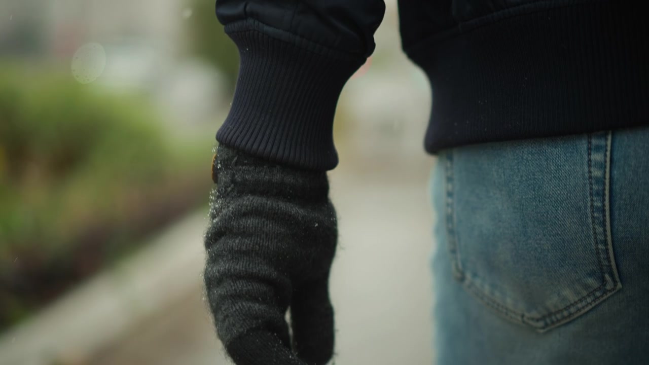 Primo piano della mano di una persona con guanto nero e jeans blu che cammina su un marciapiede bagnato con sfondo di vegetazione sfocata. Le mani guantate si muovono naturalmente mentre la persona cammina, mostrando una sfocatura di movimento costante.