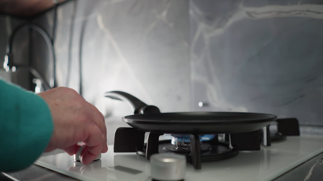 Close-up shot of elderly woman in green adjusting gas stove burner in modern kitchen. Focus on her hand turning burner knob with sleek black and white kitchen design in background