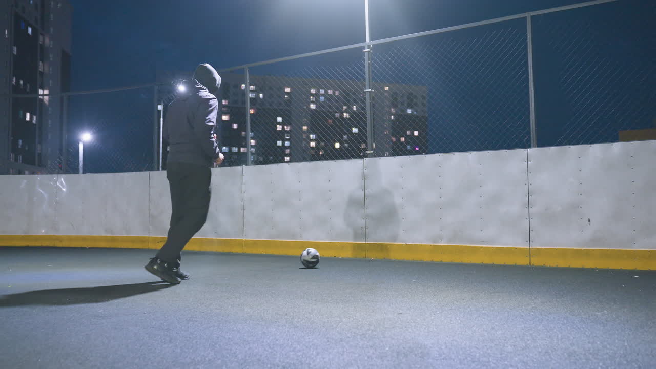 Coach kicking soccer ball against wall on illuminated urban sports field at night, with reflection visible on wall and cityscape lights in background