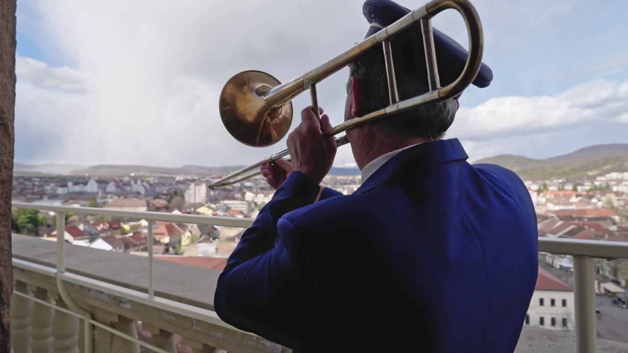 Following shot from behind shows a man in a blue uniform playing a brass trombone. The shot features an expansive background of a European town and rolling hills under a bright, cloudy sky