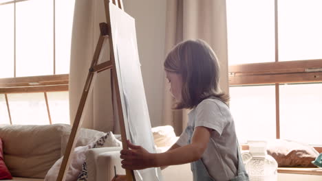 side view of a blonde girl painting her drawing with a brush on a lectern in the living room at home 1