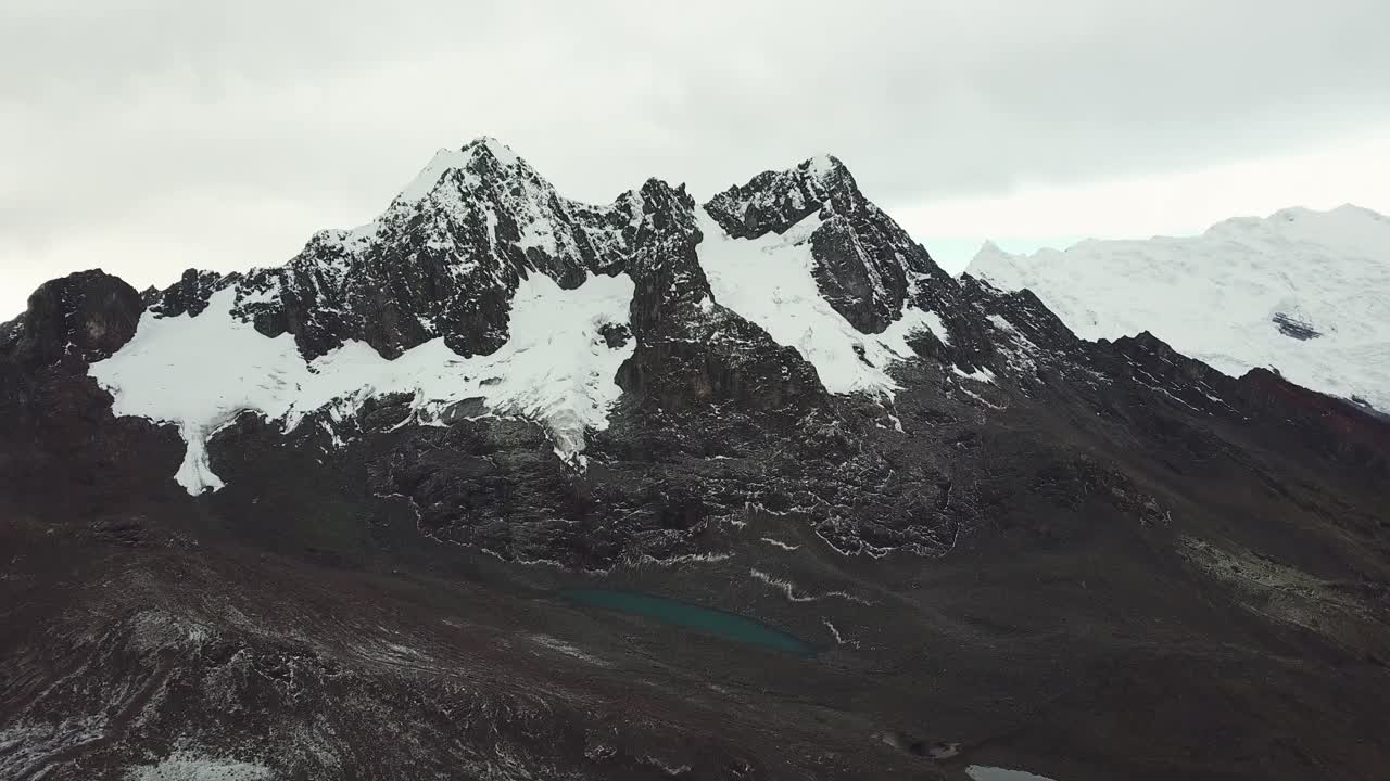 Aerial, rising, drone shot of towards rocky mountains peaks, with snow, dark, overcast day, in Peru, Andes, South America