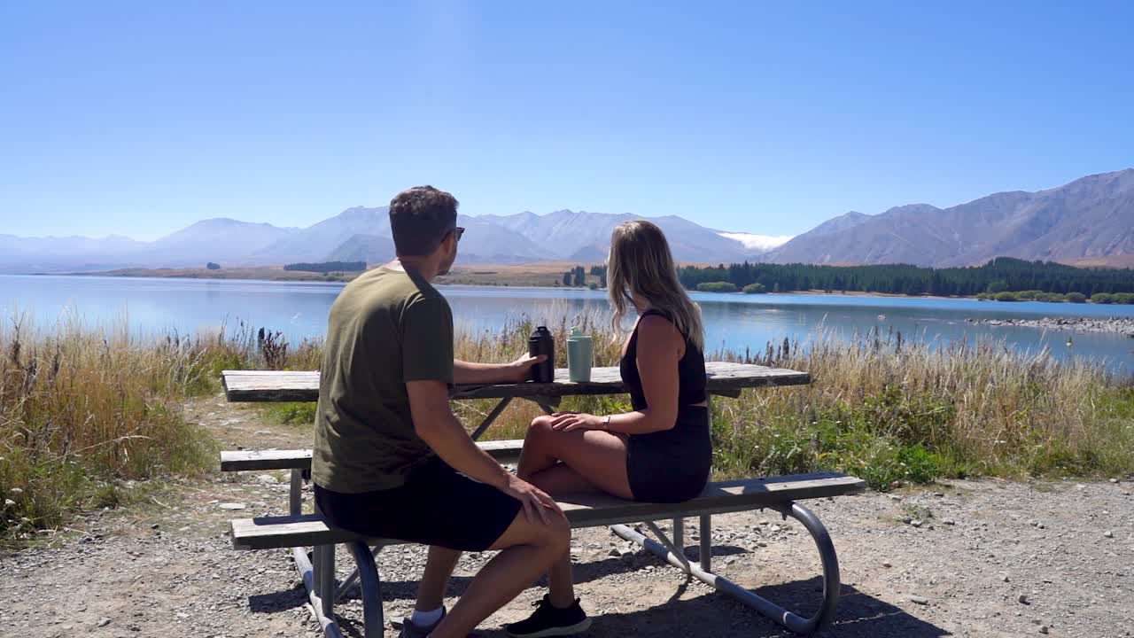 A couple sitting at a bench and drinking coffee next to Lake Tekapo in New Zealand's South Island. You can see the stunning lake and mountain scenery all around them