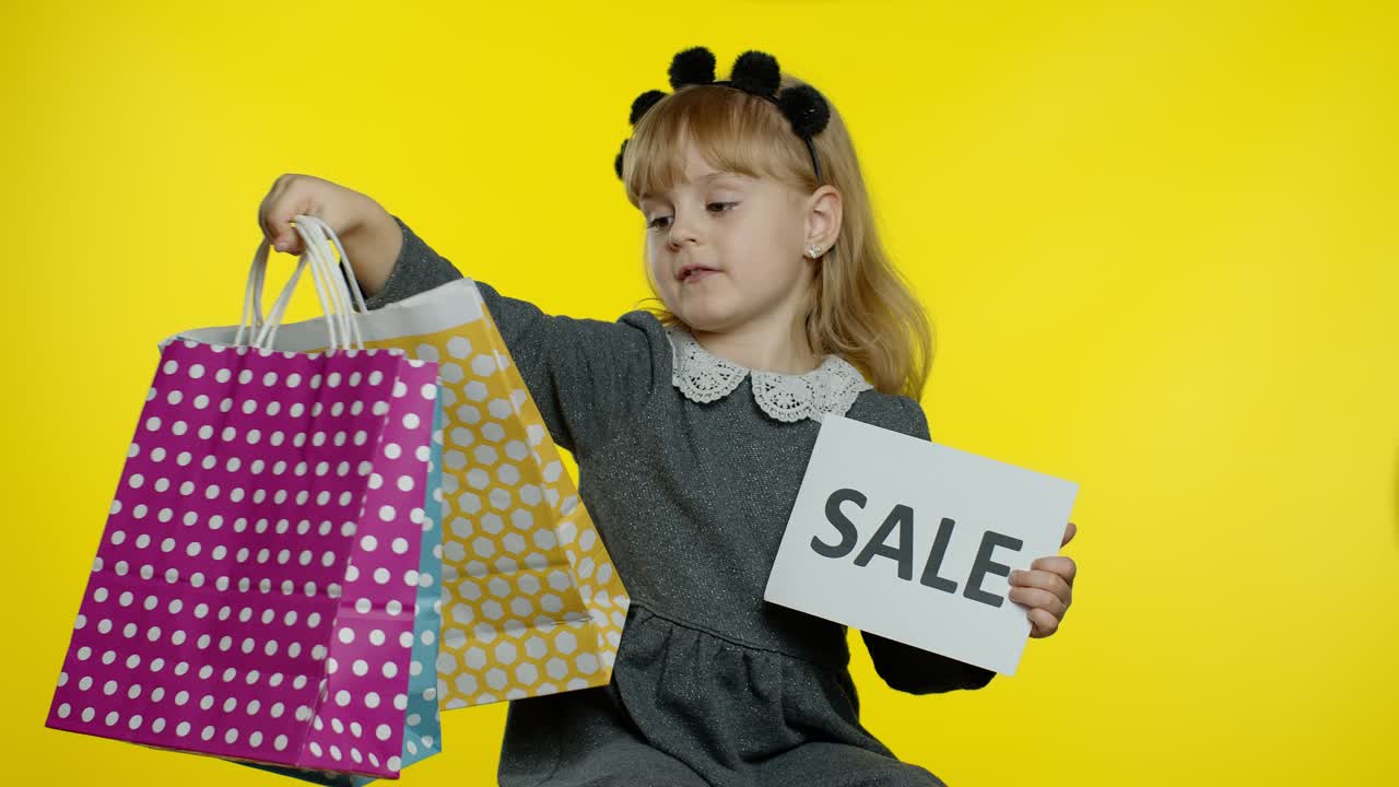 Cute joyful kid girl in shirt showing Sale word advertisement inscription banner and shopping bags