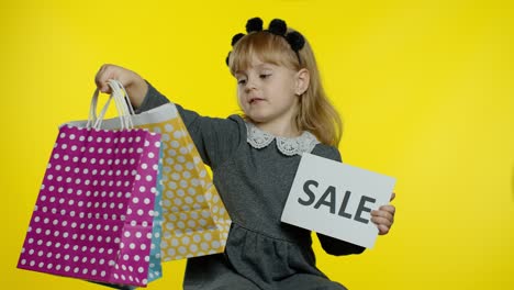 Cute-joyful-kid-girl-in-shirt-showing-Sale-word-advertisement-inscription-banner-and-shopping-bags