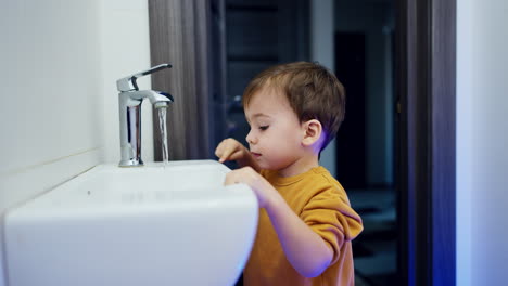 smiling cute baby stands near the sink in the bathroom. kid looks at flowing water and closes the tap.