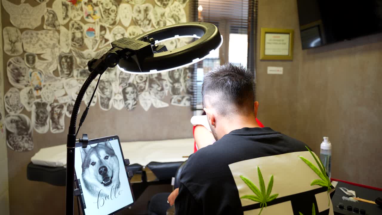 Rear shot of a tattoo artist working on a client while referencing a dog image on a tablet in the studio